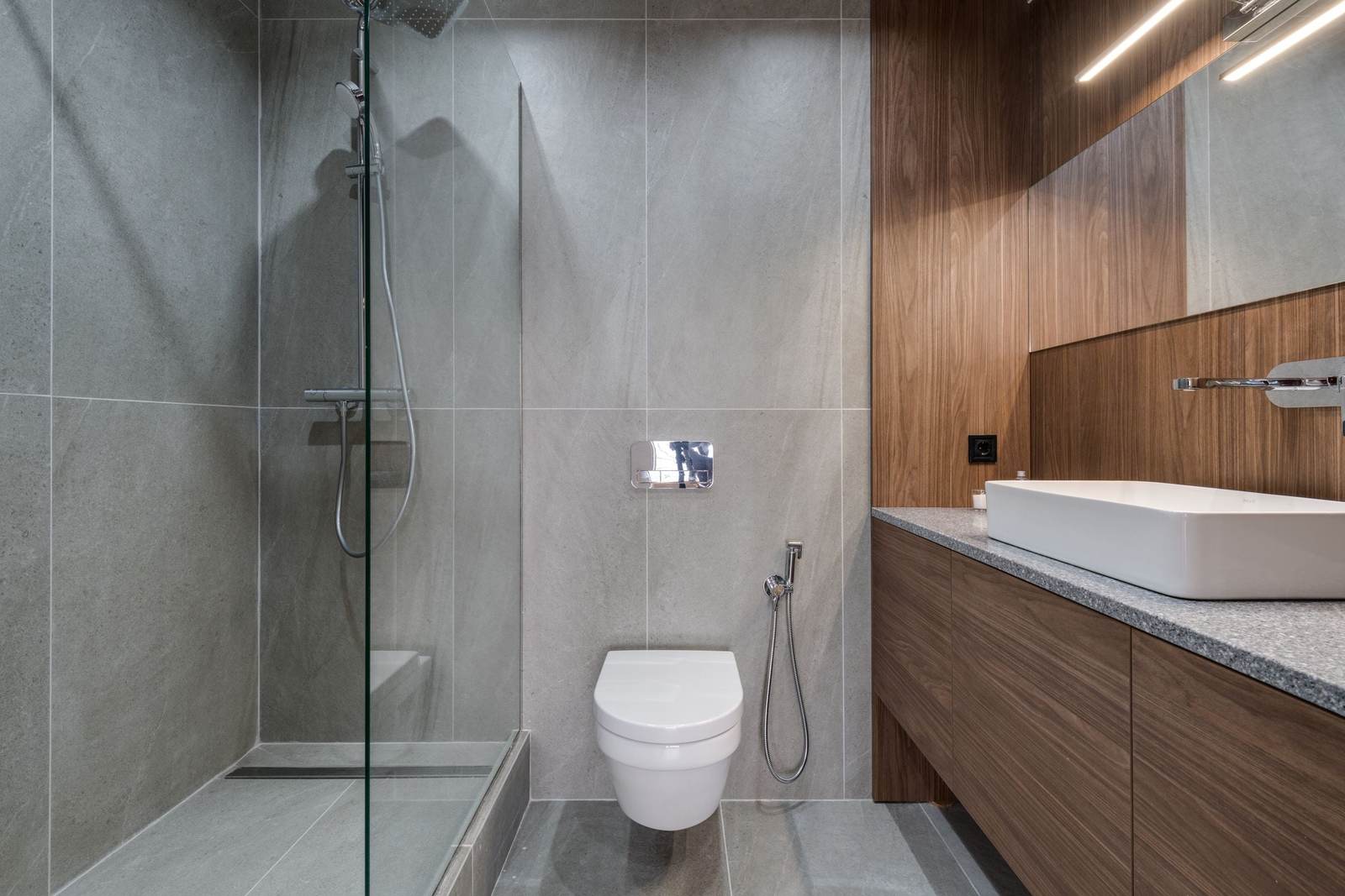 Gray porcelain shower with a low tiled threshold, a linear drain and a glass panel, beside a wall-hung toilet and a walnut vanity