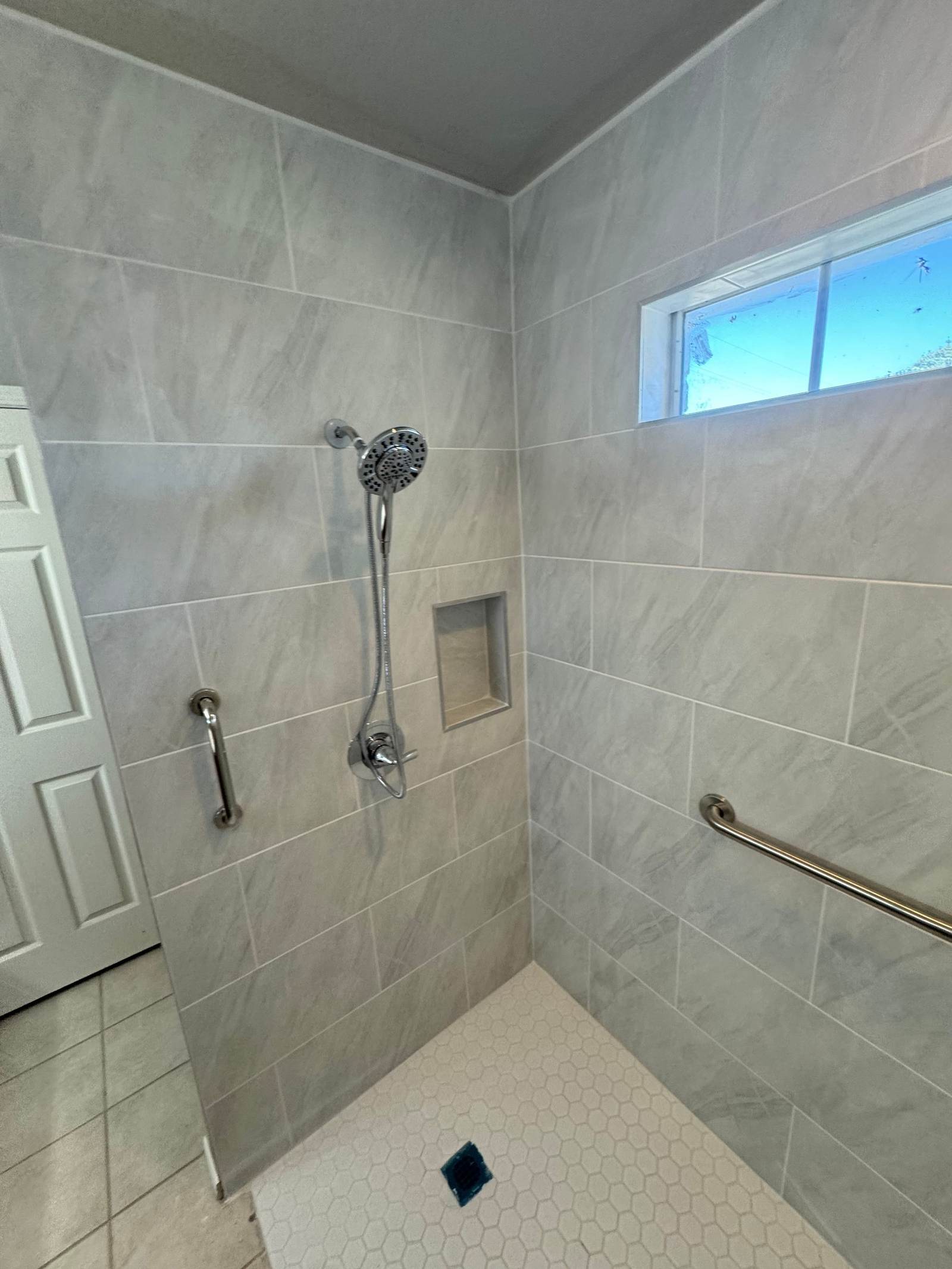 Walk-in shower with grab bars and a hand shower on a slide bar under a high window, from the roll-in shower we built for a veteran in Glenpool
