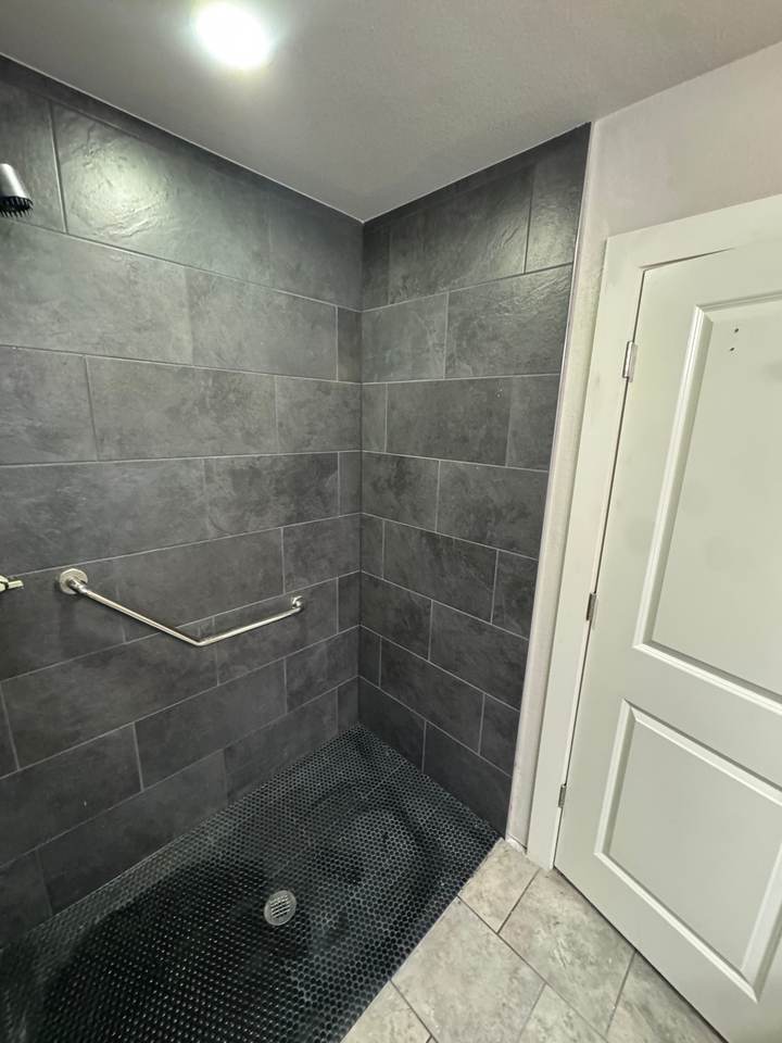 An accessible walk-in shower with dark tile, a black hex floor and a grab bar