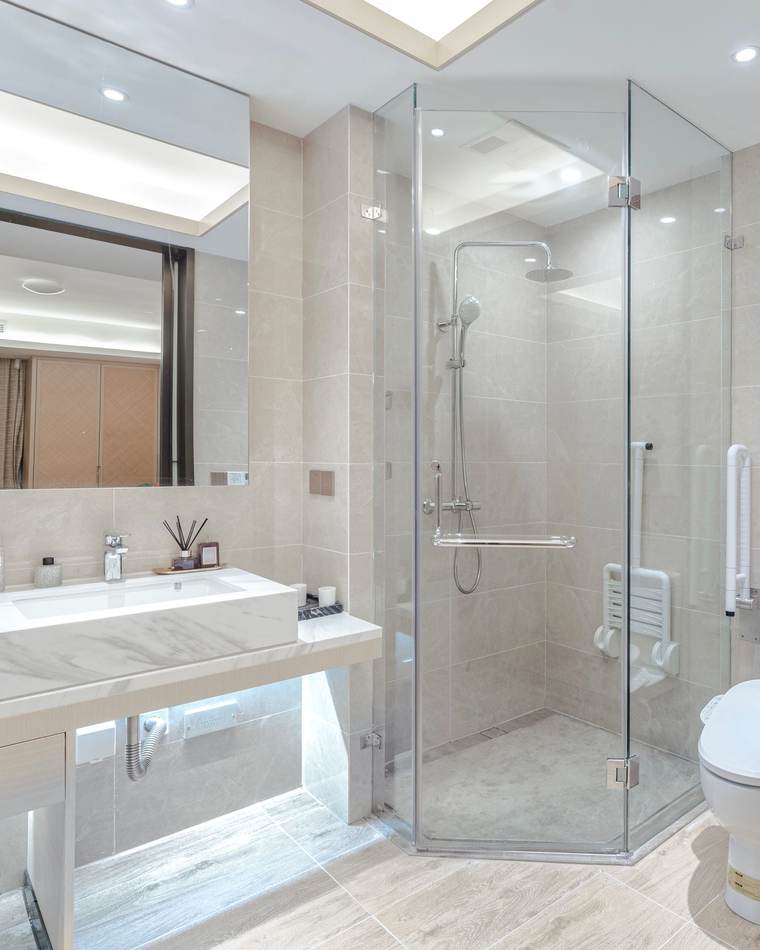 Bright, modern bathroom: a glass-walled shower with a fold-down seat, grab bars and a hand shower on a sliding rail, beside a floating marble-look vanity