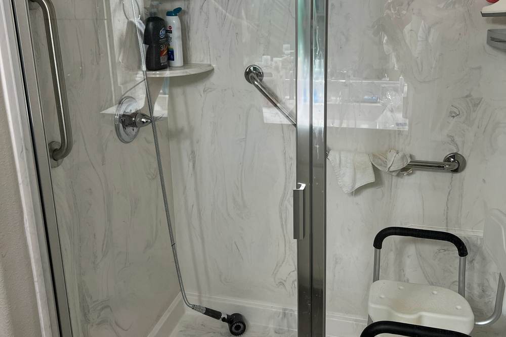 Before: a worn acrylic shower stall with a glass door, a shower chair and add-on grab bars in a Bixby home