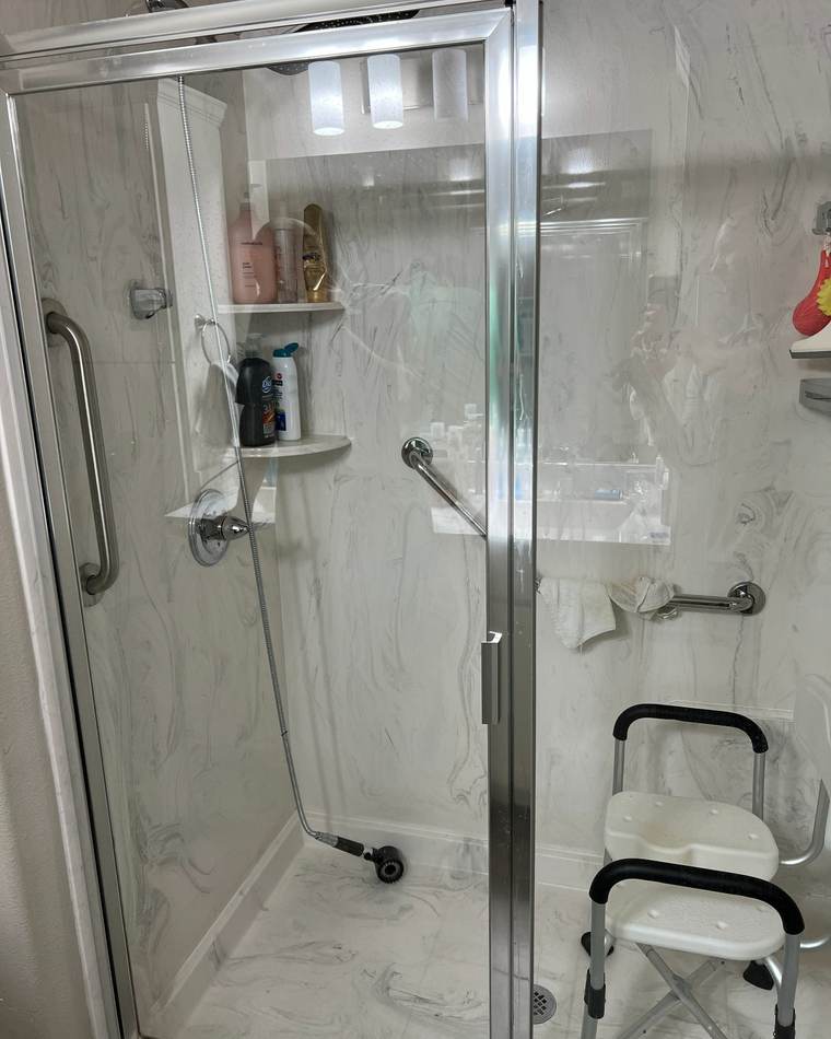 Before: an acrylic shower with a glass door, a shower chair and grab bars