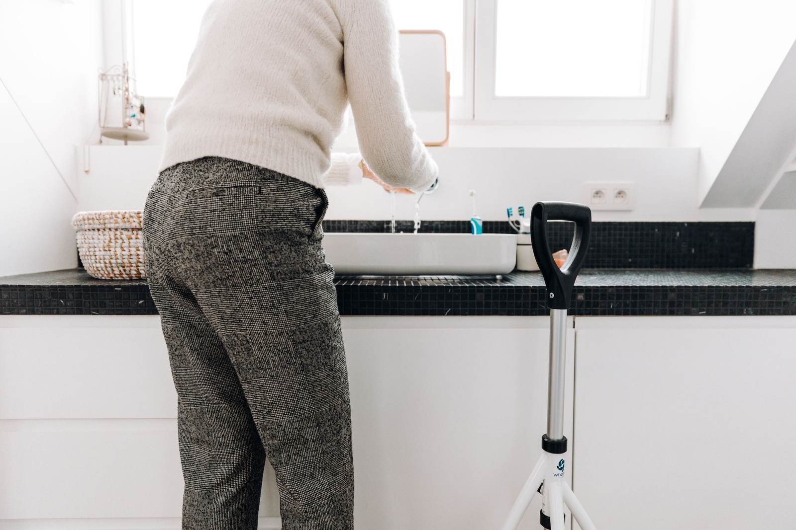 Person with a rollator standing at a white bathroom vanity with a dark countertop, under a bright window