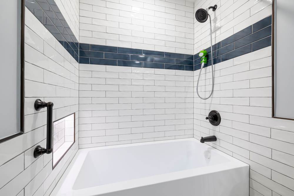 Alcove bathtub with white subway tile, a navy accent band, a black grab bar and a hand shower on a slide bar