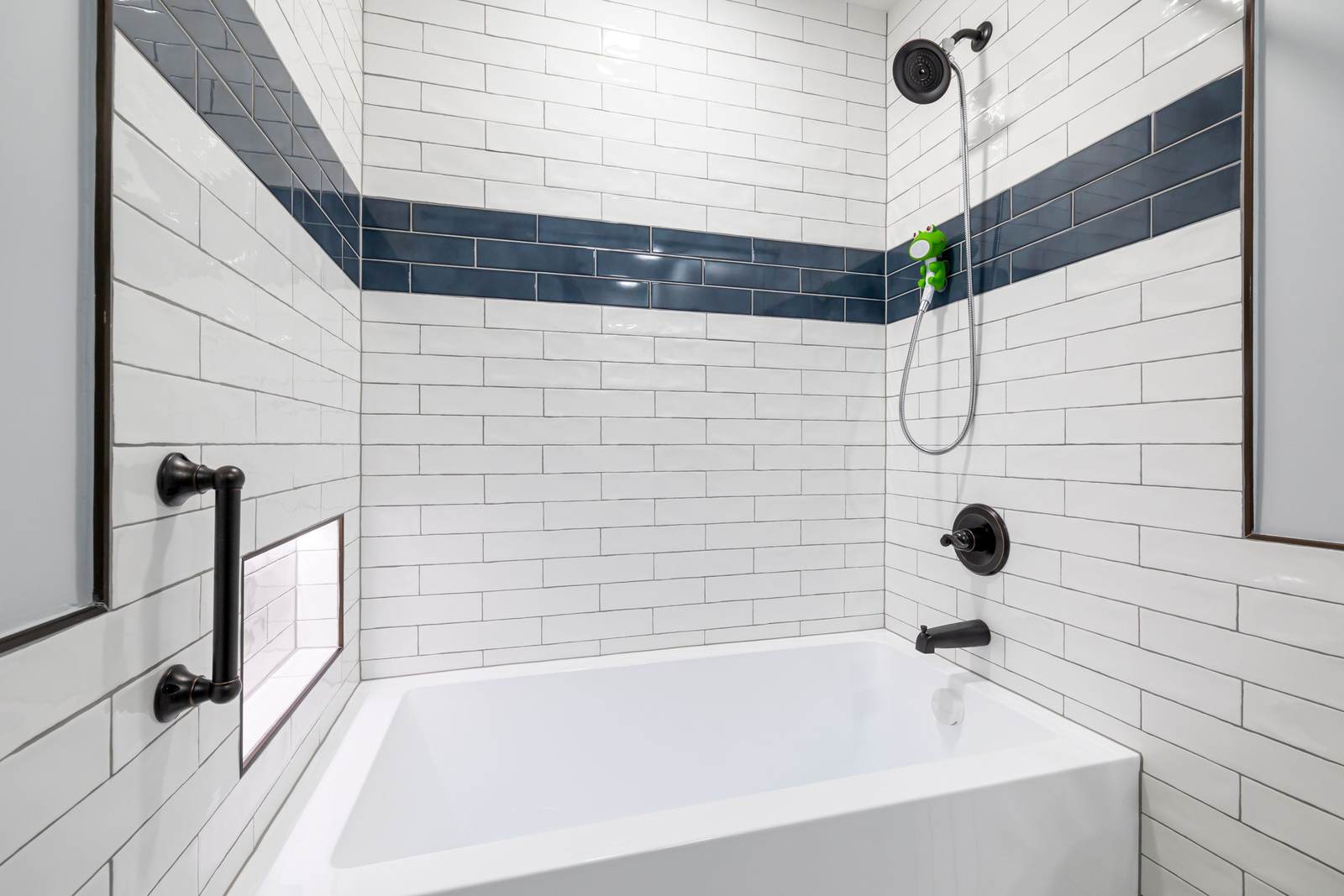 Bathtub in a white subway tile alcove with a navy accent stripe, a black grab bar and a hand shower