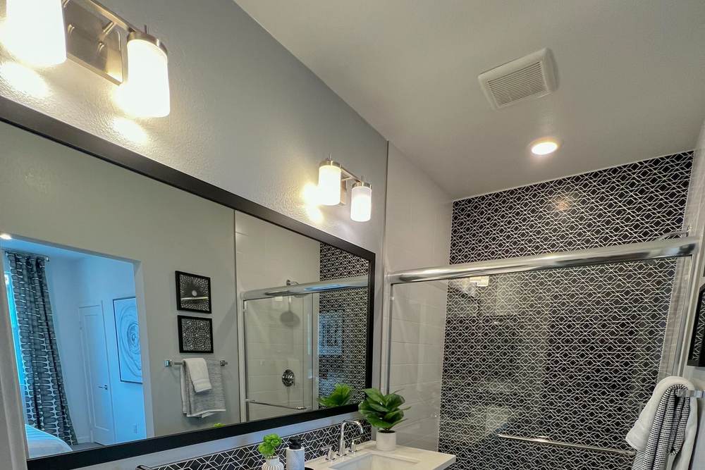 White exhaust fan grille in a bathroom ceiling between a double vanity and a tiled shower