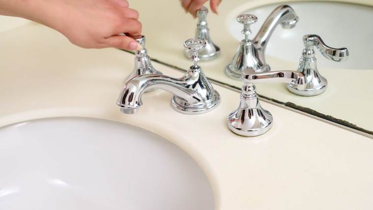 Hands fitting a chrome widespread faucet with cross handles onto a bathroom sink