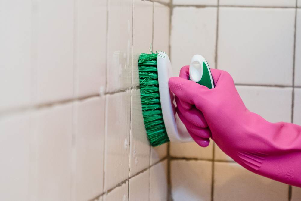 Gloved hand scrubbing mildew out of the grout where a tiled wall meets a bathtub, the kind of damage a weak fan leaves