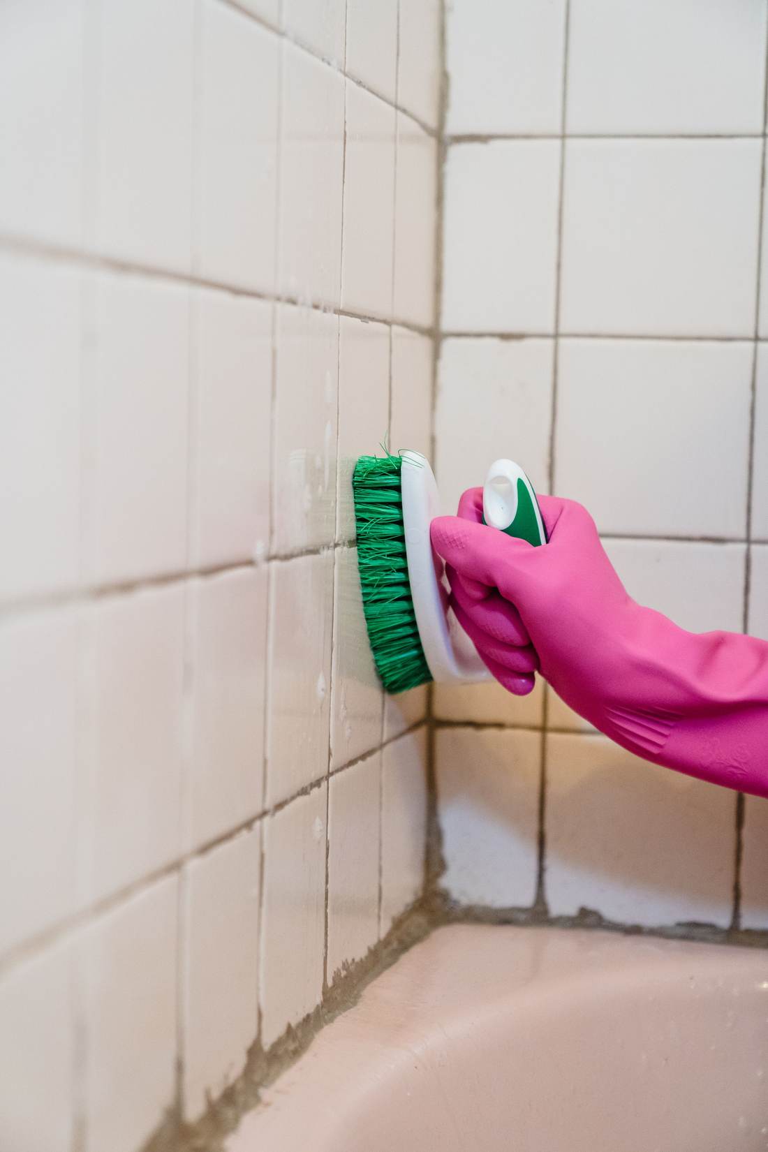 Gloved hand scrubbing white wall tile and grout with a brush