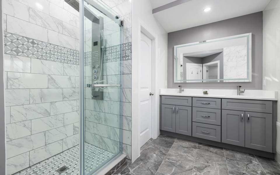 Gray double vanity with a lit mirror beside a glass walk-in shower with marble-look tile