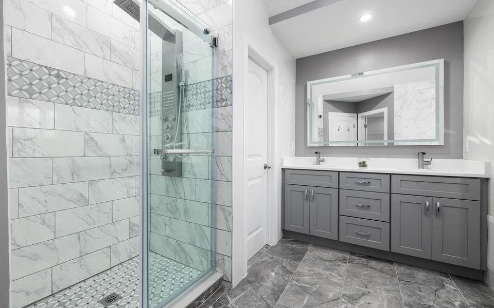 Marble-look tiled shower behind a glass door beside a gray vanity with a lit mirror