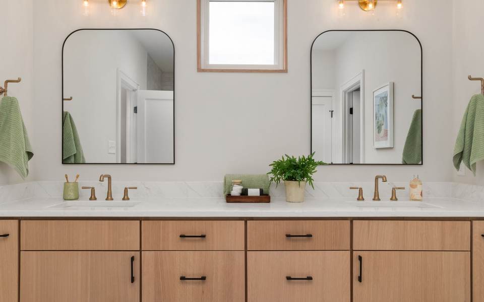 Light oak double vanity with brass faucets, black-framed mirrors and a brass light over each sink
