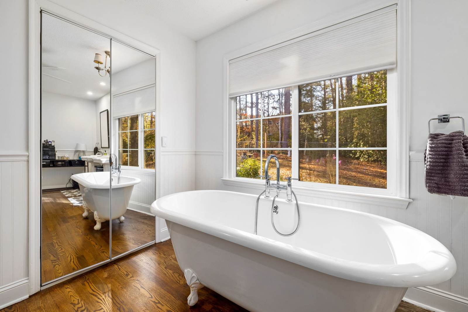 Clawfoot soaking tub under a wide window, with white wainscot walls and a wood floor