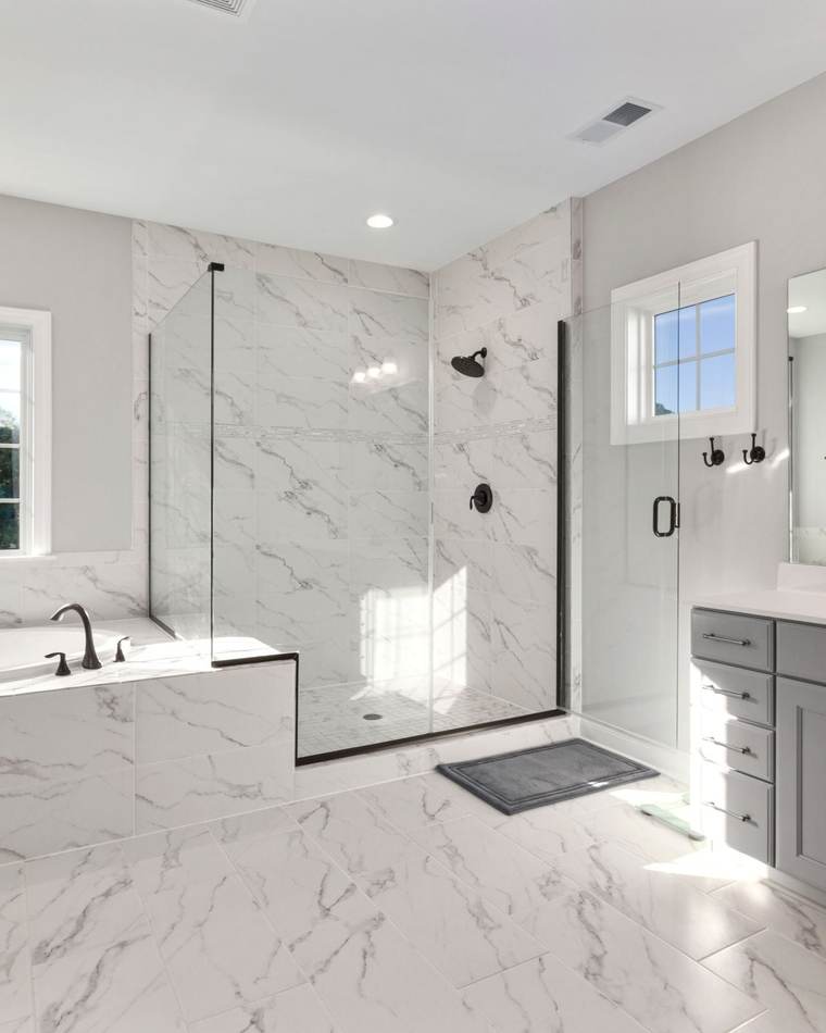 Bright primary bathroom with a frameless glass shower, marble-look tile and a tiled drop-in tub under a window