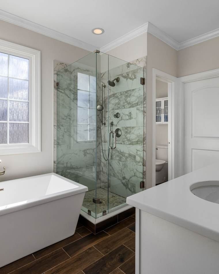 Bright primary bathroom with a freestanding soaking tub under a window, a marble-look glass shower and a white vanity with brushed nickel faucets