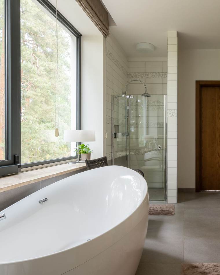 Bright bathroom with a white freestanding tub under a tall window looking out on pine trees, a glass shower and a wood vanity