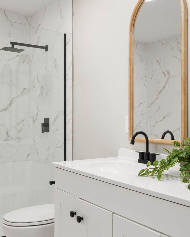 Updated bathroom with a marble-look tiled shower behind a glass panel, a white double vanity, matte black faucets and arched wood mirrors
