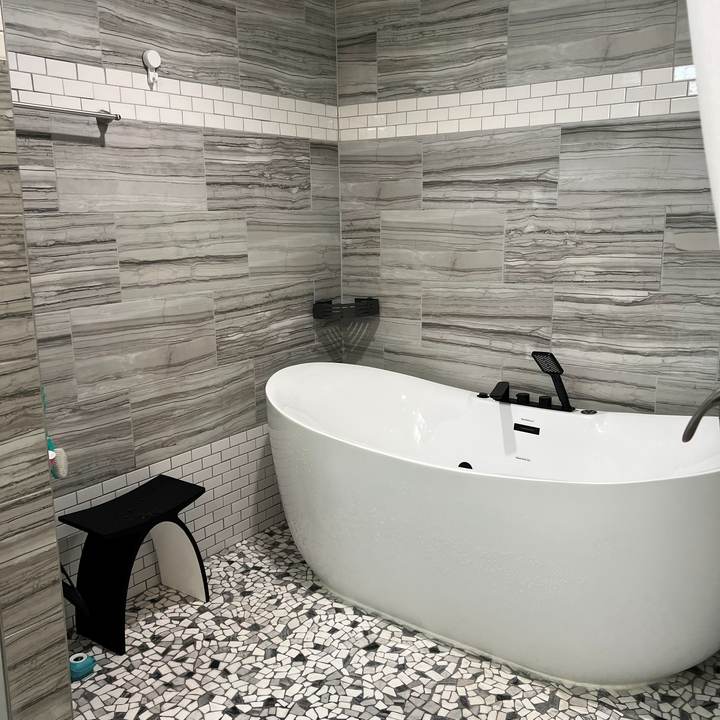 A freestanding tub and wood bench in a gray tiled wet room with a subway tile band
