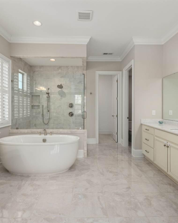 Bright primary bathroom with a large marble-look porcelain tile floor, a freestanding tub in front of a glass shower, and a cream vanity