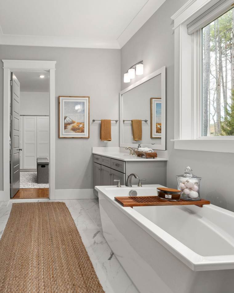 Bright primary bathroom with a freestanding tub under a window onto tall trees, a glass-walled marble-look shower and a gray vanity