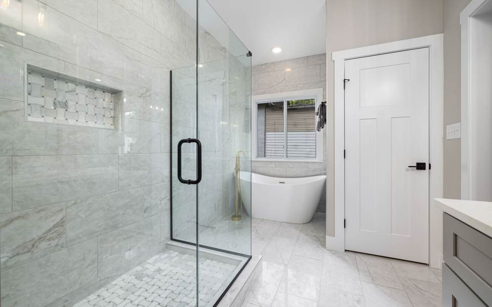 Marble-look walk-in shower behind a glass door, with a freestanding tub under a window beyond it