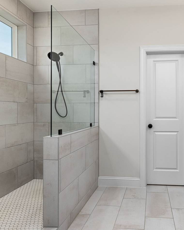 Walk-in shower behind a tiled half wall with a glass panel, beside a navy vanity with a round wood mirror