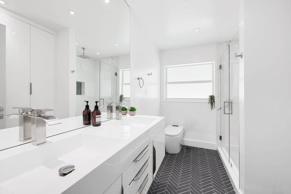 Narrow white bathroom with a long vanity, a dark herringbone tile floor and a hinged glass shower door at the far end