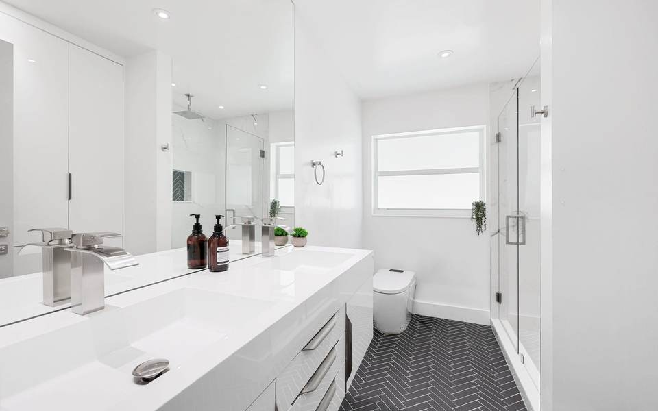 Long white vanity with a one-piece top and brushed nickel faucets beside a frameless glass shower, over a dark herringbone floor