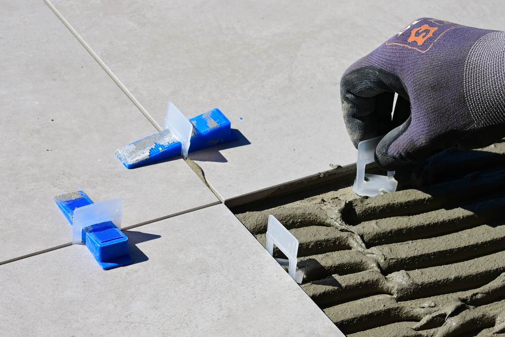 Gloved hand setting tile leveling clips between large gray porcelain floor tiles laid in fresh thin-set