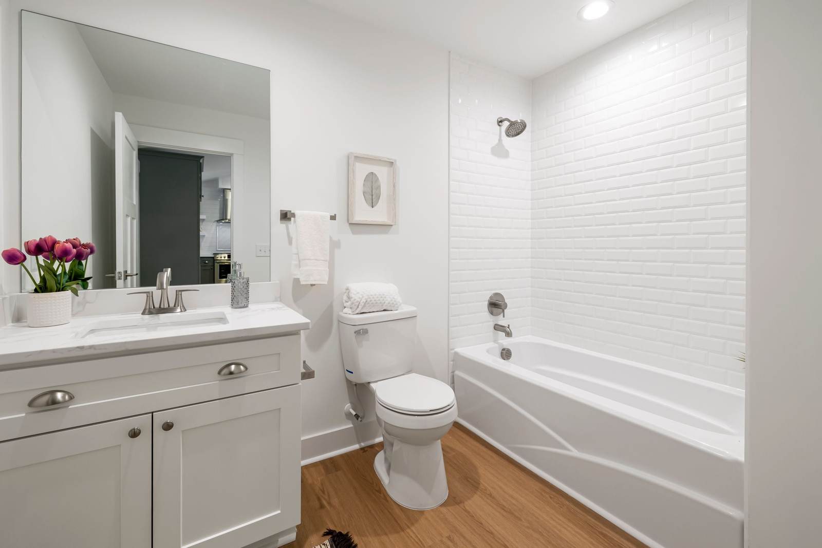 New white alcove bathtub with a beveled white subway tile surround and brushed nickel trim, beside a white shaker vanity