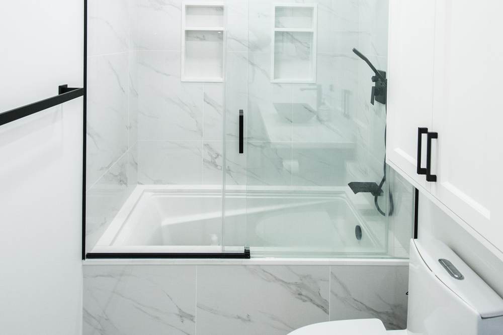 New soaking tub behind a sliding glass door, with marble-look tile walls, two niches and matte black fixtures