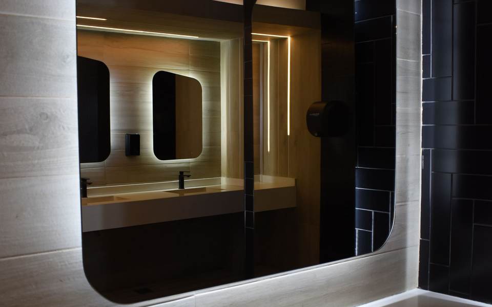 Restroom with backlit rounded mirrors over a floating counter and dark wall tile