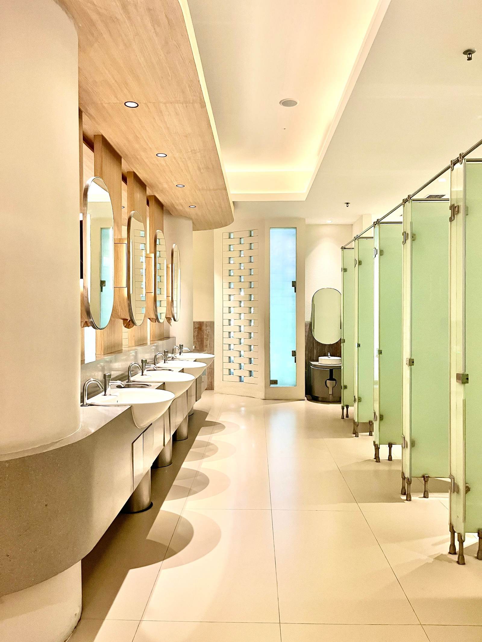 Bright commercial restroom with a long row of sinks, round mirrors, and frosted partitions