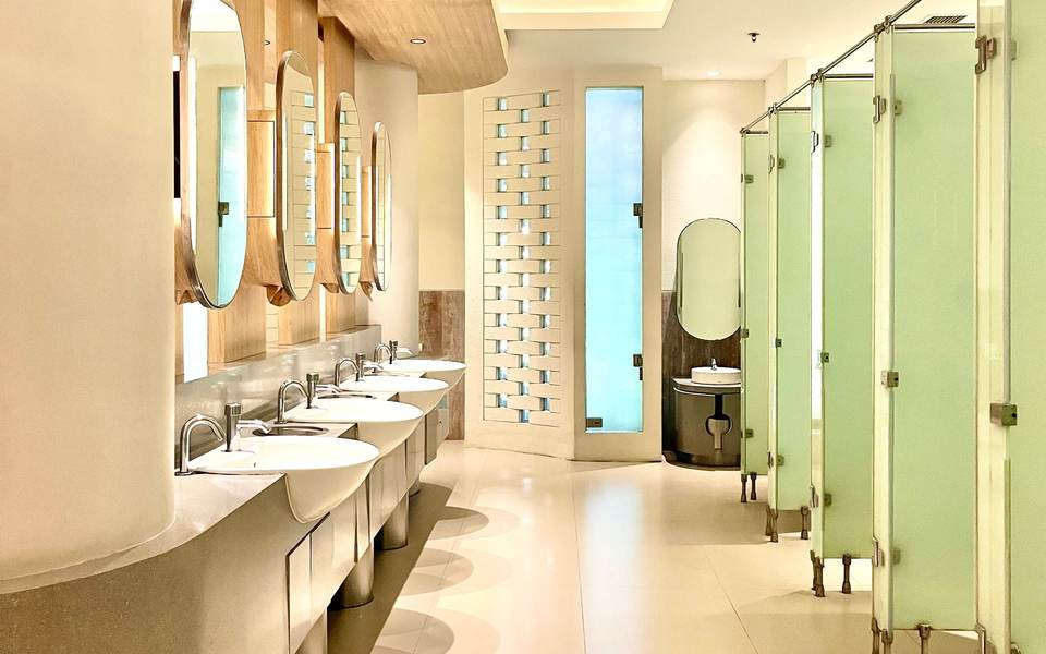 Bright commercial restroom with a row of sinks under round mirrors across from pale green toilet partitions