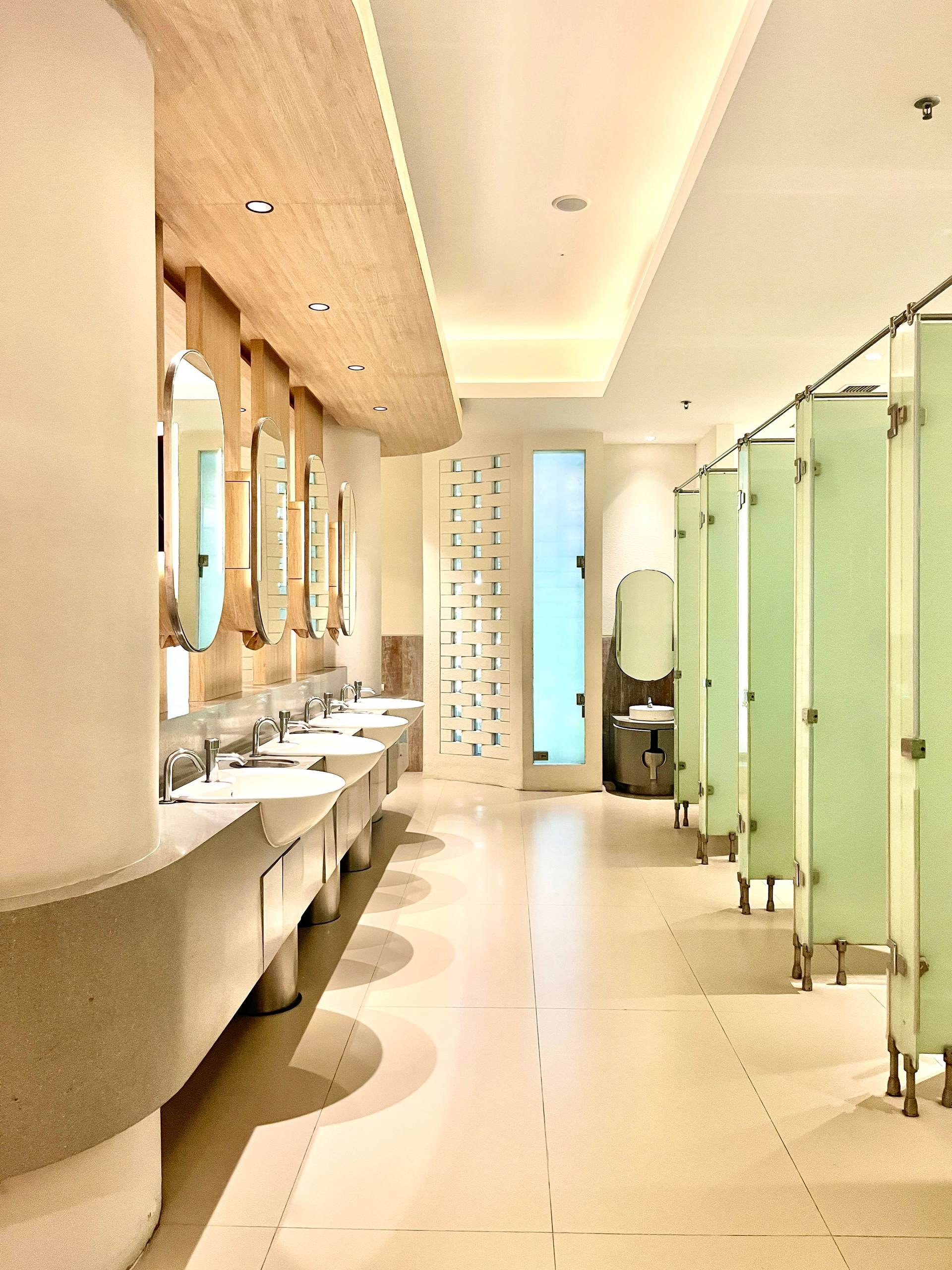 Bright commercial restroom with a long row of sinks, round mirrors, and frosted toilet partitions