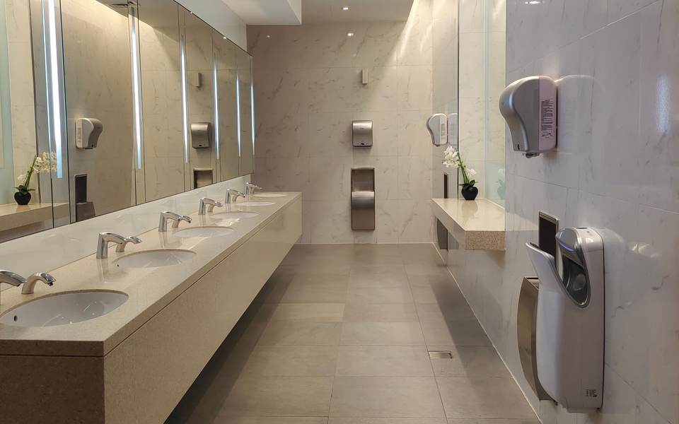 Long restroom counter in pale stone with a row of undermount sinks, lit mirrors and hand dryers on the wall