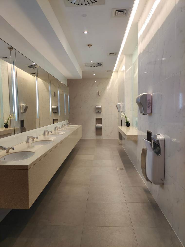 Long commercial restroom with a light tile floor, a stone counter and wall-mounted hand dryers