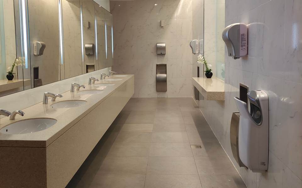 Long commercial restroom with a floating stone counter, a row of sinks, backlit mirrors and wall-mounted hand dryers