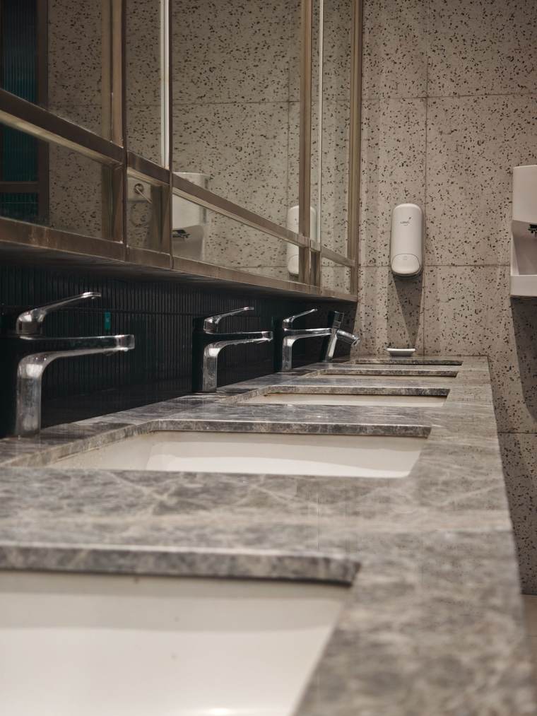 Row of stainless steel faucets over undermount sinks in a stone restroom counter