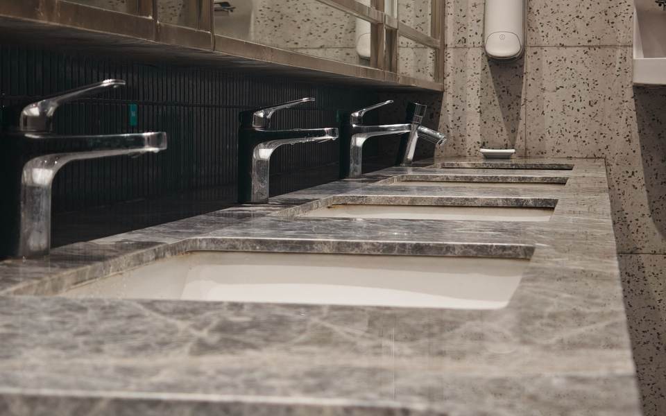 Row of undermount sinks in a stone restroom counter with stainless faucets under a long mirror