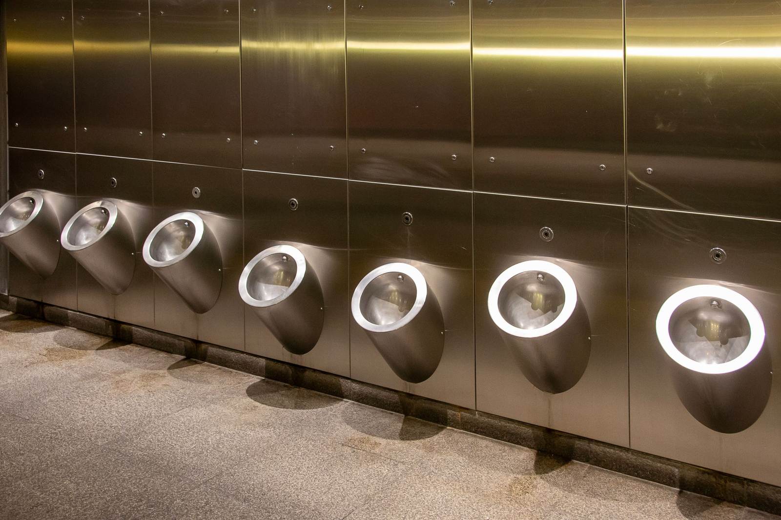 Row of stainless steel urinals with lit rims along a brushed metal wall