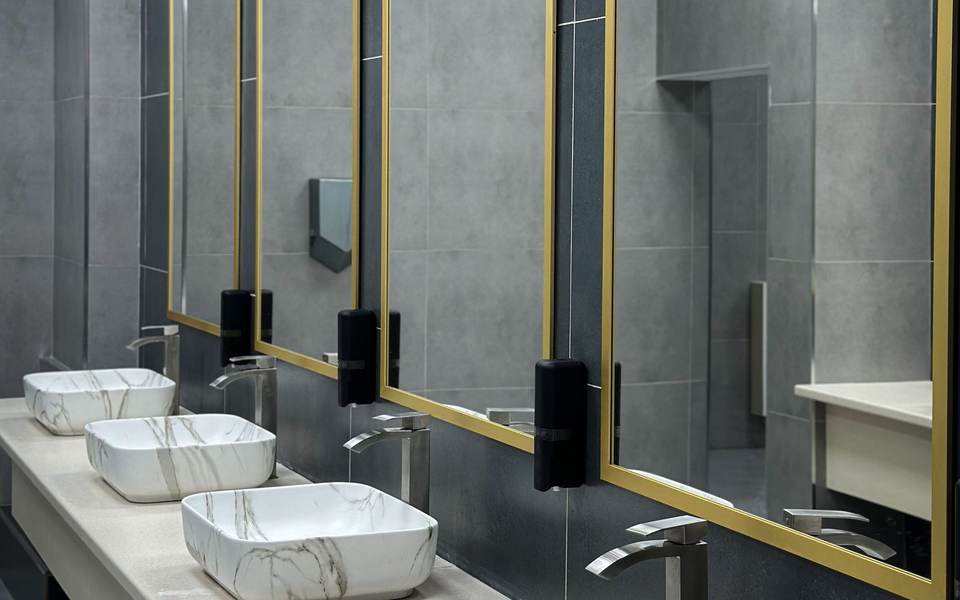 Restroom with gray tile walls, brass-framed mirrors and a row of marble-look vessel sinks on a long counter