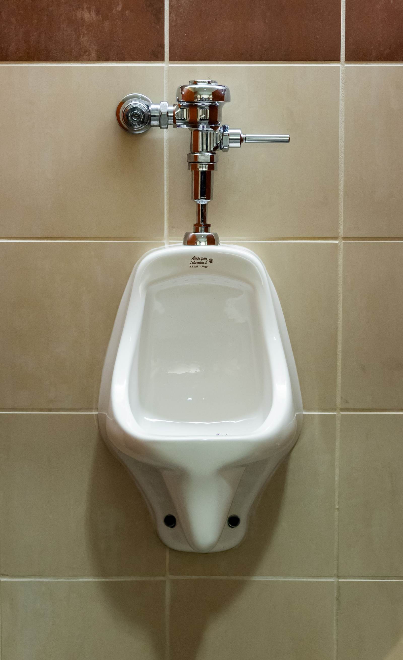 Wall-hung urinal with a chrome manual flush valve on a beige tile wall