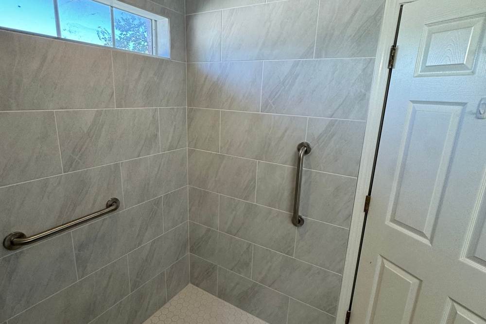 Curbless roll-in shower with grab bars and a white hex floor under a transom window, built for a veteran in Glenpool