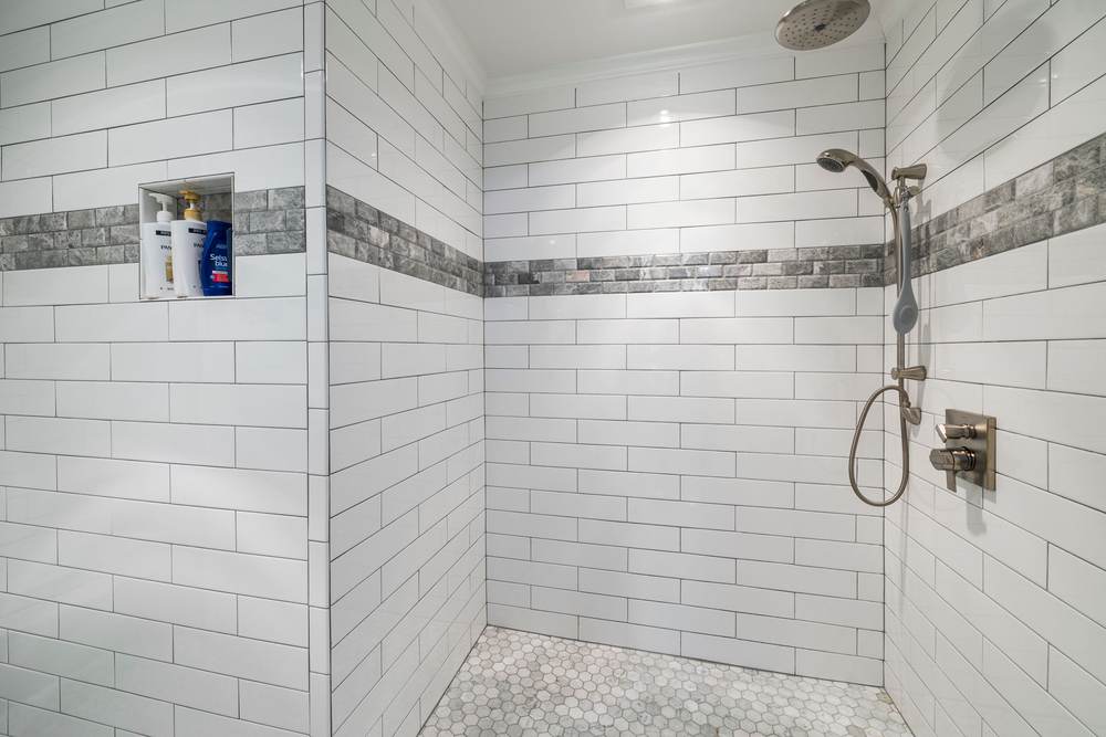 Open walk-in shower with white subway tile, a gray accent band, a hand shower on a slide bar and a tiled niche