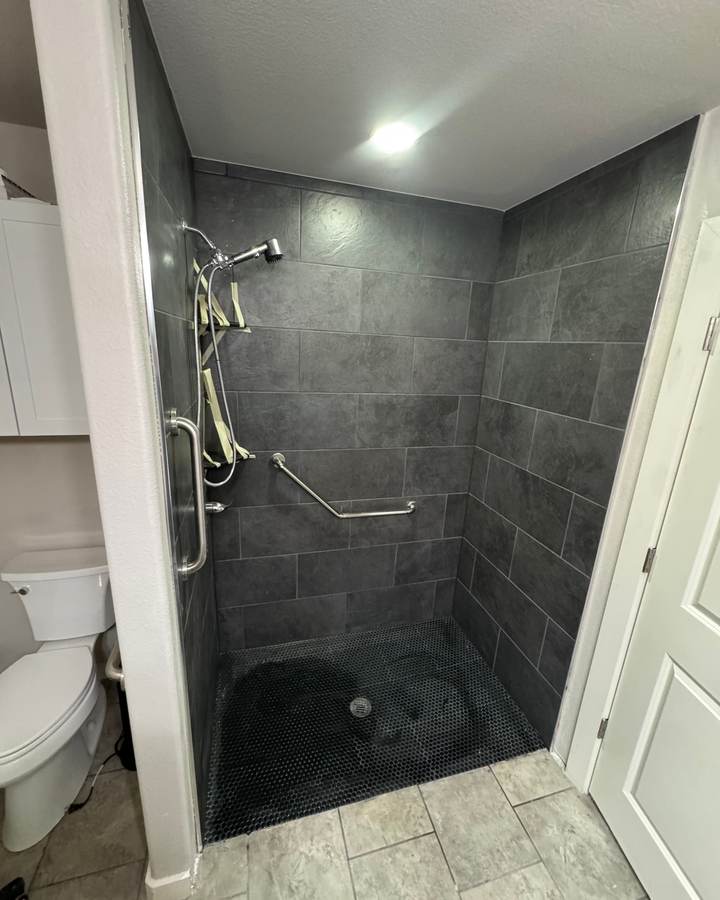 A curbless shower with slate-look tile, a dark mosaic floor and grab bars beside the toilet