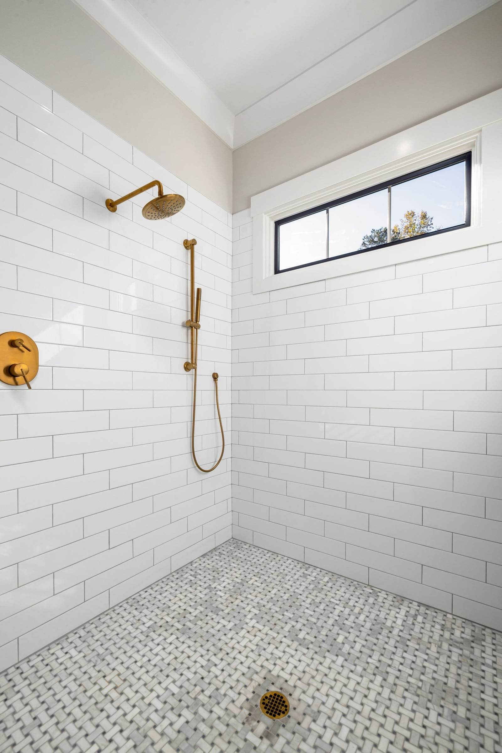Custom walk-in shower with white subway tile, a marble basketweave floor, brass shower fixtures and a transom window above the tile