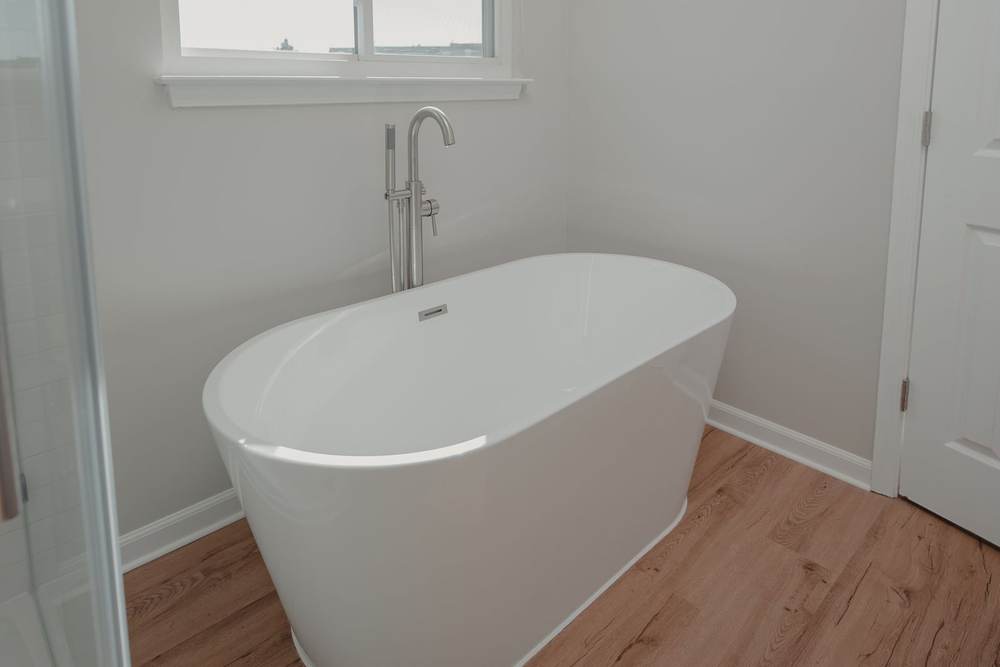 Oval freestanding tub with a floor-mounted filler under a window, set out from the walls on a wood-look floor