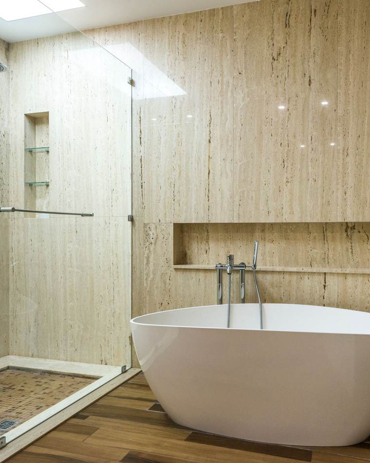 Freestanding soaking tub with a wall-mounted filler beside a frameless glass shower with a rain head, travertine-look wall tile and a wood-look floor