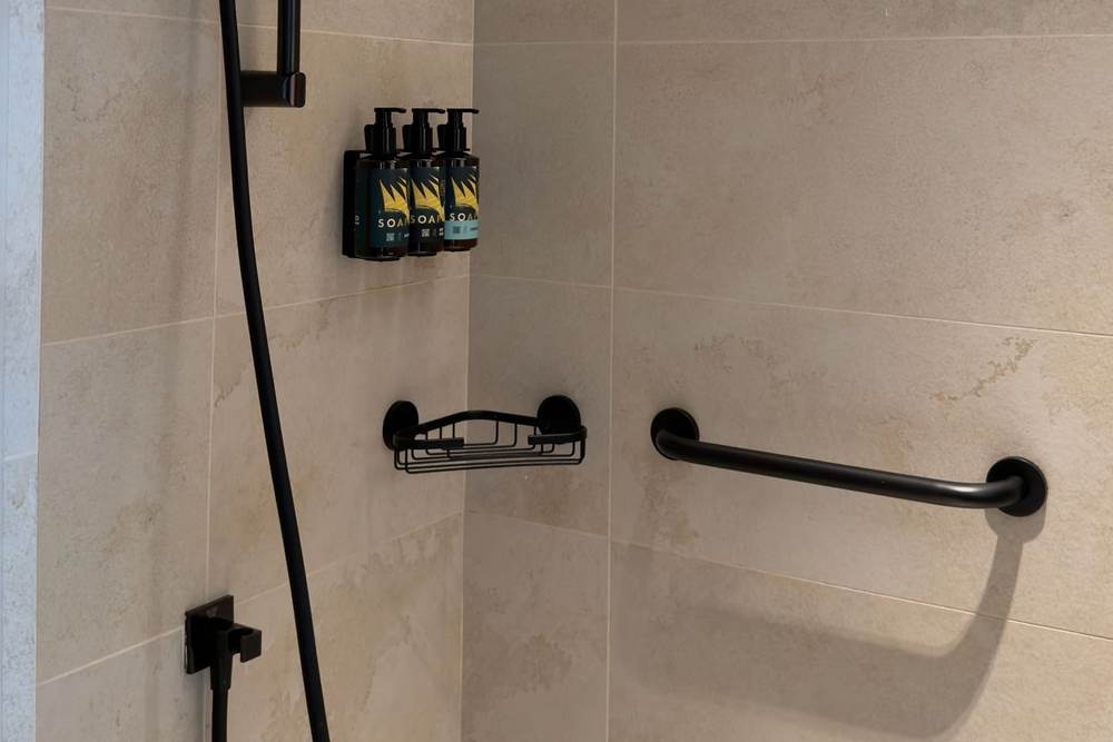 Matte black grab bar on beige stone-look shower tile beside a matching black hand shower on a slide bar